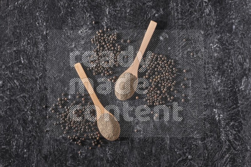 2 wooden spoons full of black pepper powder and black pepper beads spread on a textured black flooring