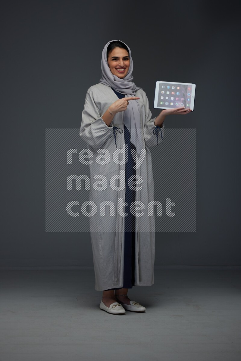 A Saudi woman wearing a light gray Abaya and head scarf standing and showing the phone's screen eye level on a grey background