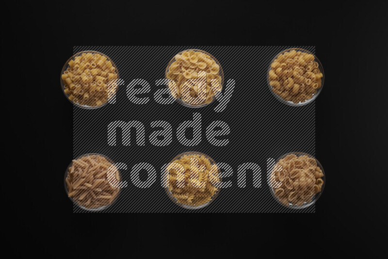 Different pasta types in 6 glass bowls on black background