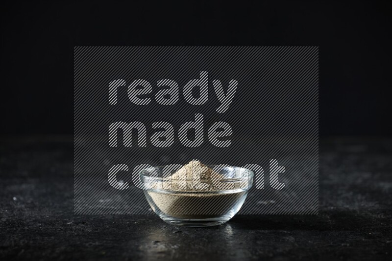 A glass bowl full of cardamom powder on textured black flooring