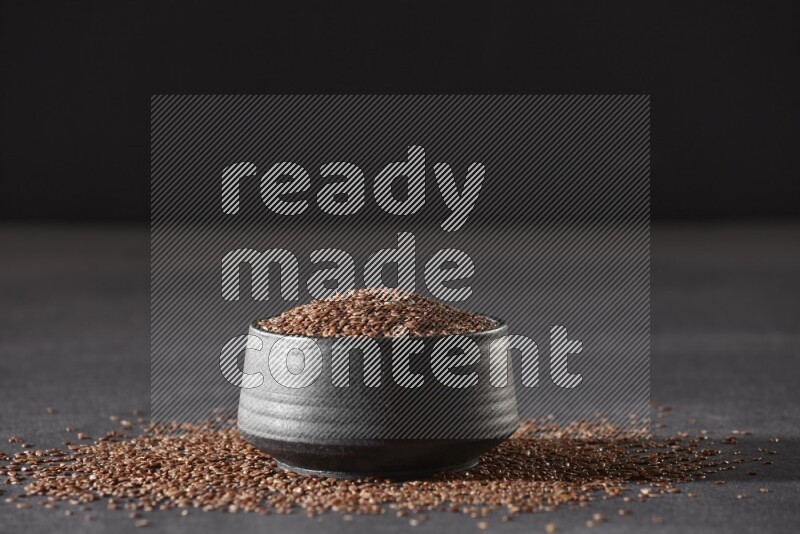 A black pottery bowl full of flaxseeds surrounded by the seeds on a black flooring