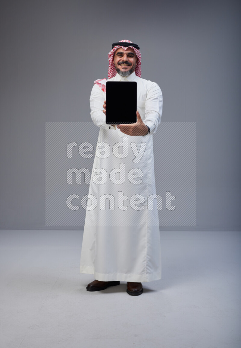 Saudi man Wearing Thob and red Shomag standing showing tablet to camera on Gray background
