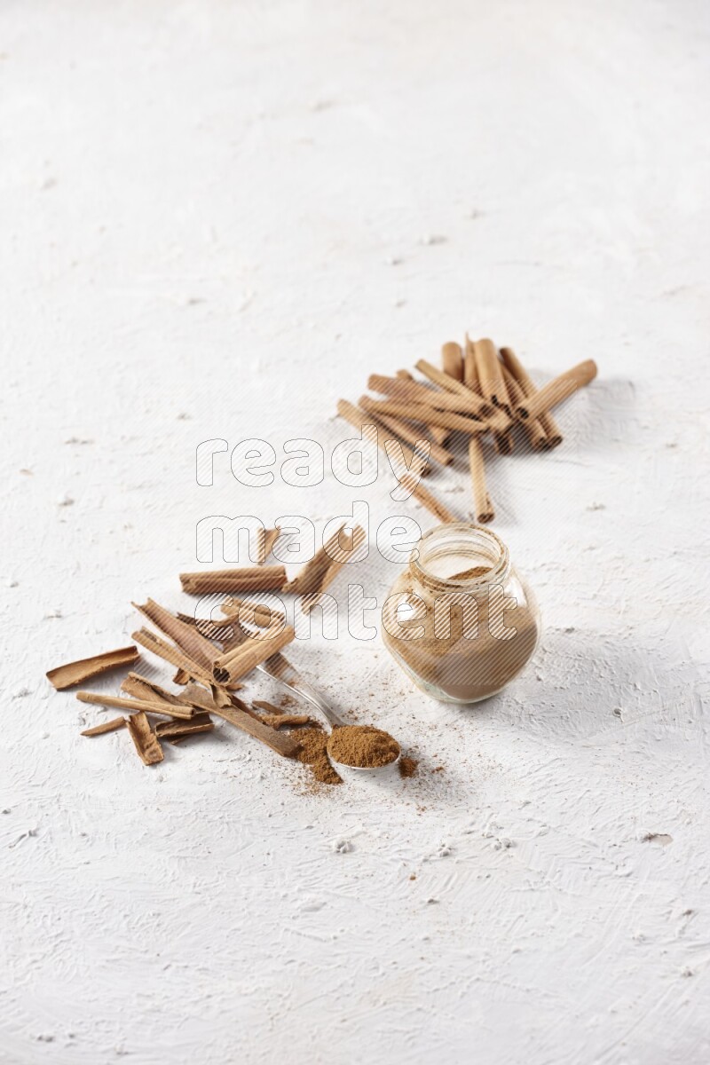 Herbal glass jar full cinnamon powder and a metal spoon surrounded by cinnamon sticks on a white background