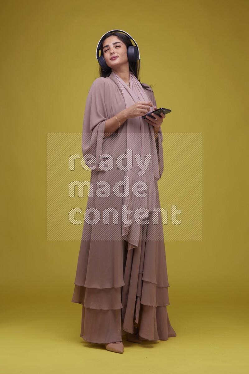A Saudi woman Listing to Music on a Yellow Background wearing Brown Abaya