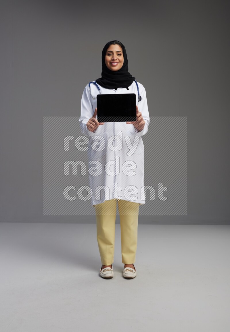 Saudi woman wearing lab coat with stethoscope standing showing tablet to camera with sign in the back on Gray background