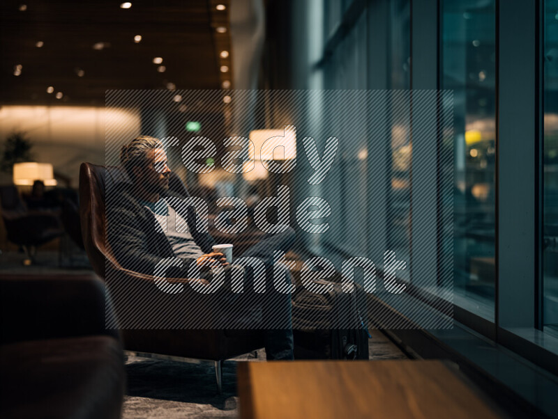 A solo traveler sits comfortably in a premium airport lounge, holding a cup of coffee while gazing through floor-to-ceiling windows at the airside view. The scene captures a calm pre-flight pause common in modern travel routines, where personal reflection and mental preparation take place before departure. The refined lounge setting, soft lighting, and organized luggage reflect efficiency, comfort, and the transitional nature of contemporary business and long-haul travel, resonating with the rhythm of international journeys through major regional hubs.يجلس مسافر منفرد في صالة مطار مميزة، ممسكًا بفنجان قهوة وهو ينظر عبر النوافذ الزجاجية الواسعة نحو ساحة الطائرات. تجسد الصورة لحظة هدوء وتأمل تسبق الرحلة، وهي جزء مألوف من روتين السفر الحديث حيث يهيئ المسافر نفسه ذهنيًا قبل الانطلاق. تعكس أجواء الصالة الراقية، والإضاءة الناعمة، وترتيب الأمتعة معاني الراحة والكفاءة والطابع الانتقالي للسفر المعاصر، بما يتماشى مع إيقاع الرحلات الدولية عبر المراكز الإقليمية الكبرى.
