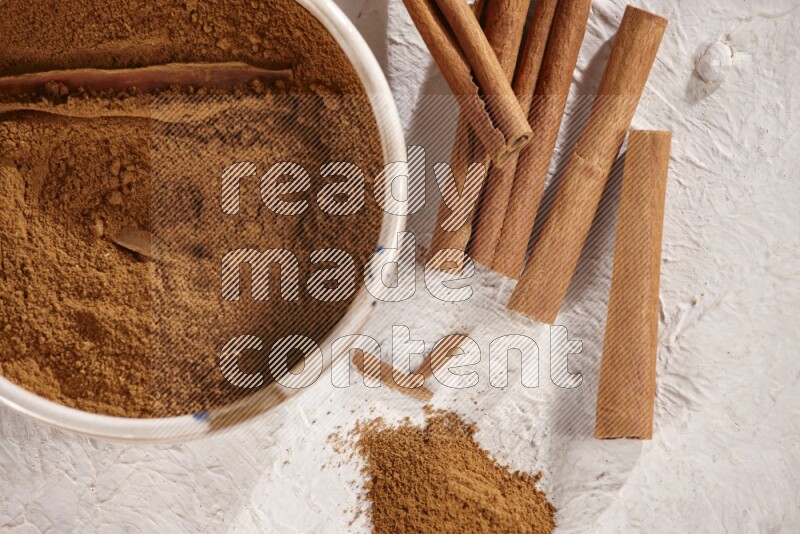 Ceramic bowl full of cinnamon powder with cinnamon sticks on the side on white background