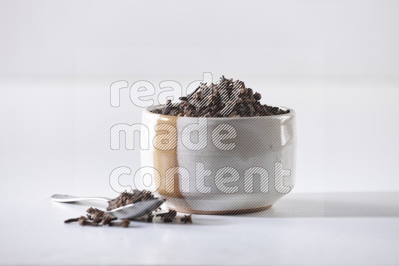A beige ceramic bowl full of cloves and a metal spoon next to it on a white flooring