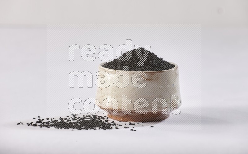 A beige pottery bowl full of black seeds and more seeds spread on a white flooring