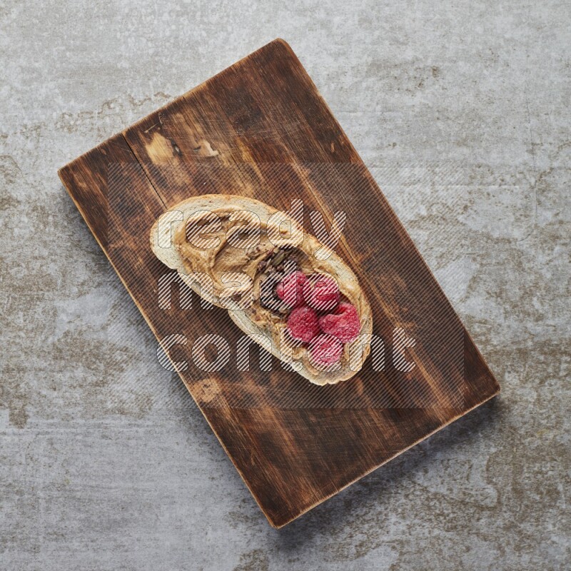 open faced peanut butter sandwich with raspberries and chocolate granula on a grey textured background