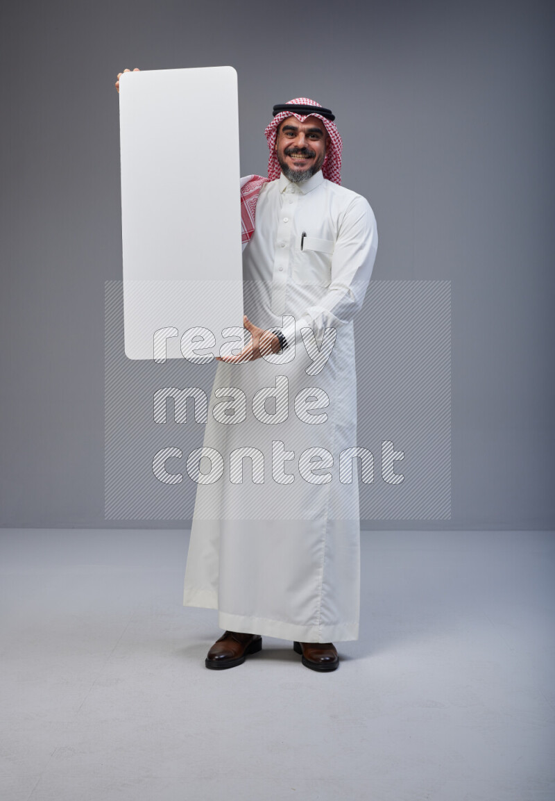 Saudi man Wearing Thob and red Shomag standing holding board on Gray background