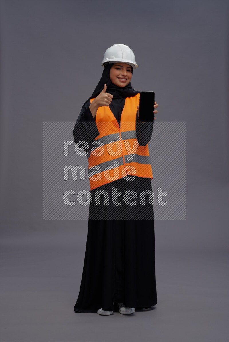 Saudi woman wearing Abaya with engineer vest standing showing phone to camera on gray background