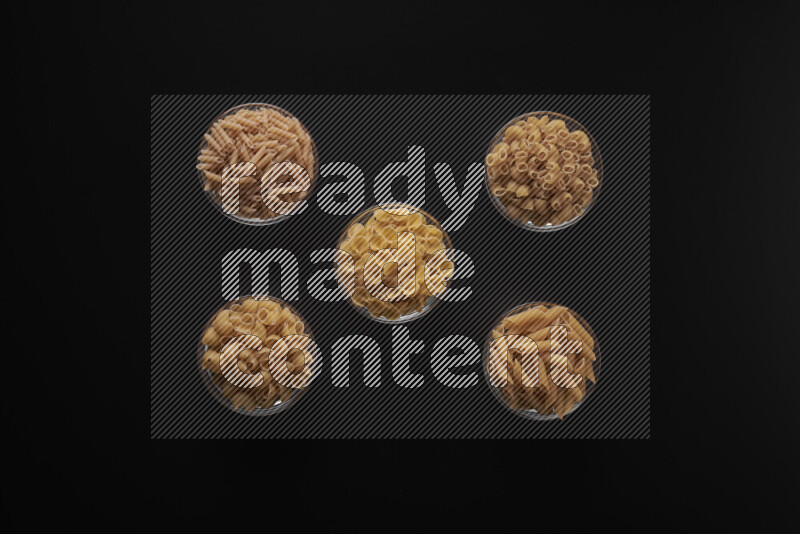 Different pasta types in 5 glass bowls on black background