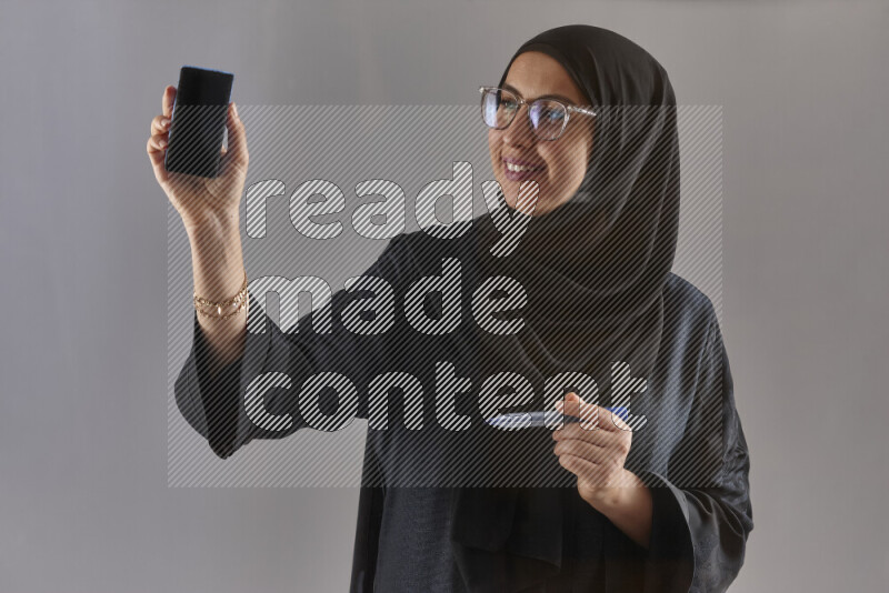 Woman in black abaya, hijab and glasses holding a marker pen to write on a transparent board with different reactions and expressions on grey background