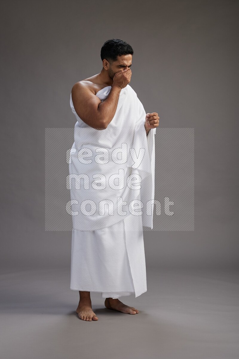 A man wearing Ehram standing showing how to sneeze on gray background