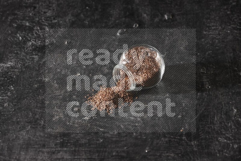 A glass spice jar full of flaxseeds flipped and seeds spread out on a textured black flooring