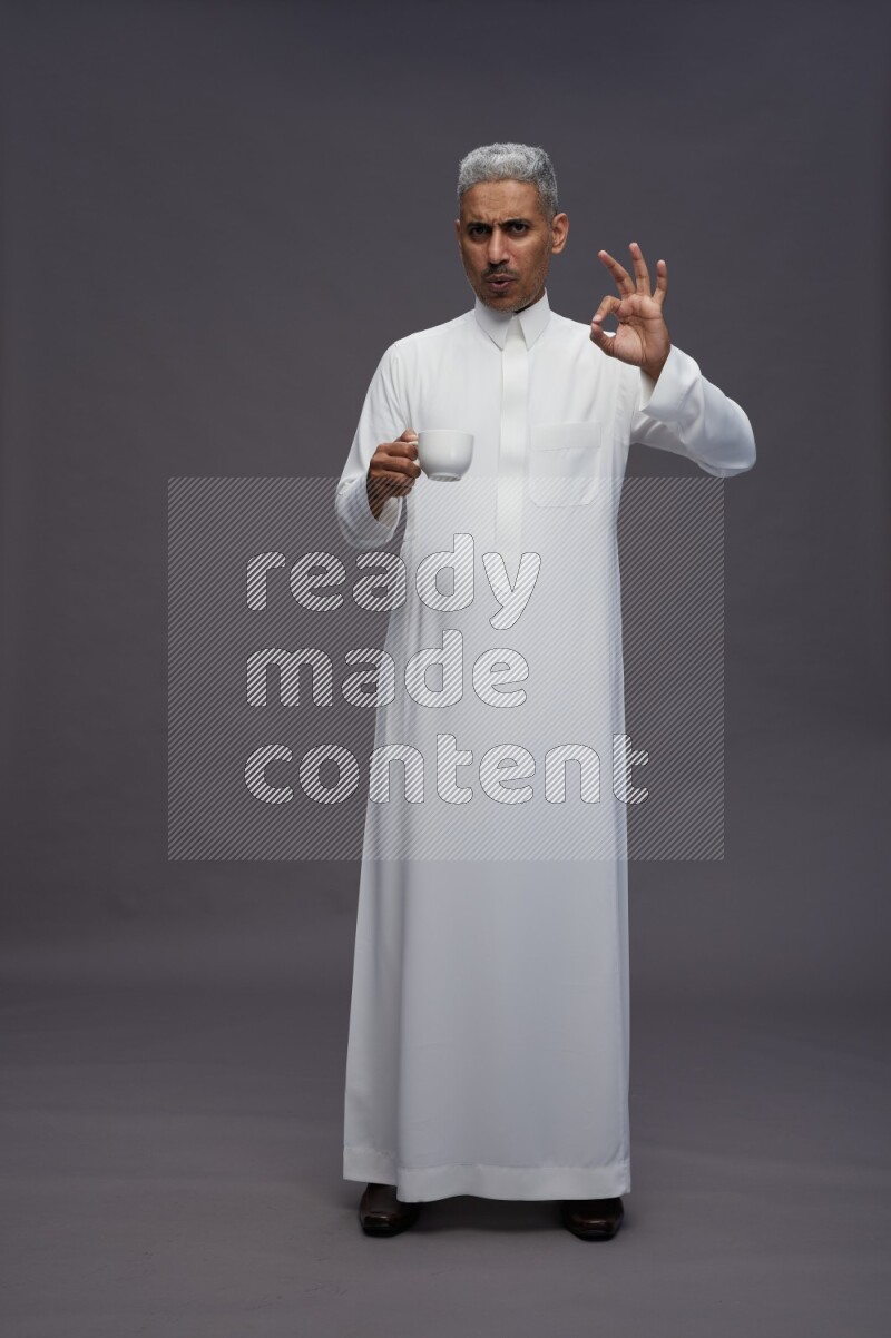 Saudi man wearing thob standing holding mug on gray background