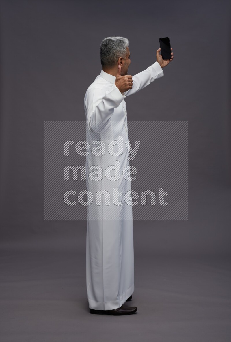 Saudi man wearing thob standing taking selfie on gray background