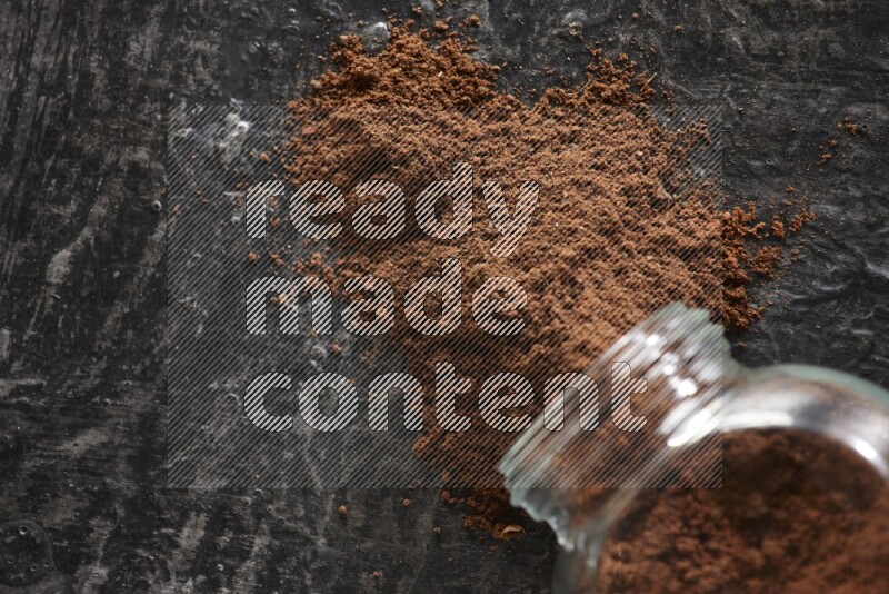 A flipped glass spice jar full of cloves powder and powder came out of it on textured black flooring