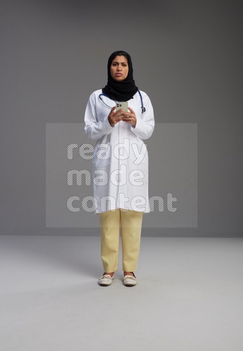 Saudi woman wearing lab coat with stethoscope standing texting on phone on Gray background