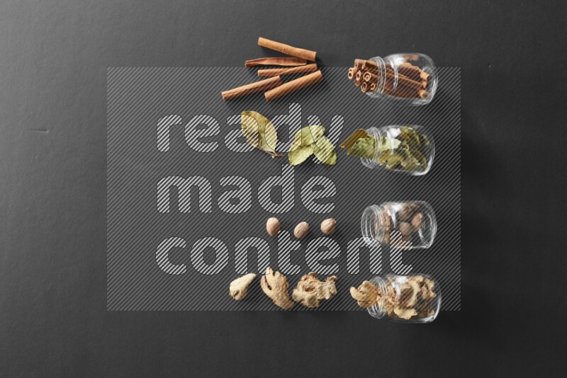 4 glass spice jars full of laurel bay leaves, ginger, cinnamon and nutmeg on black flooring