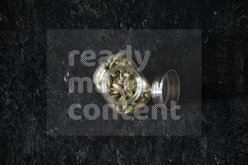 A glass spice jar full of cardamom seeds on textured black flooring