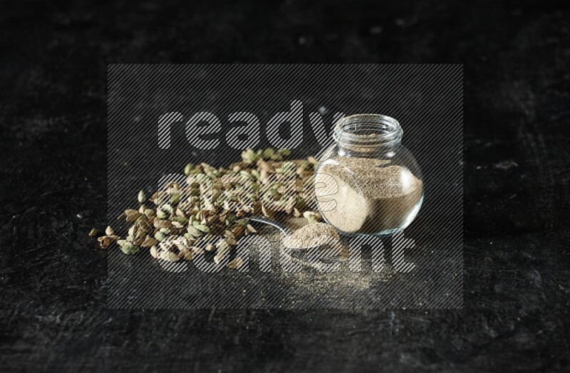 A glass spice jar and a metal spoon full of cardamom powder and cardamom seeds spreaded on textured black flooring