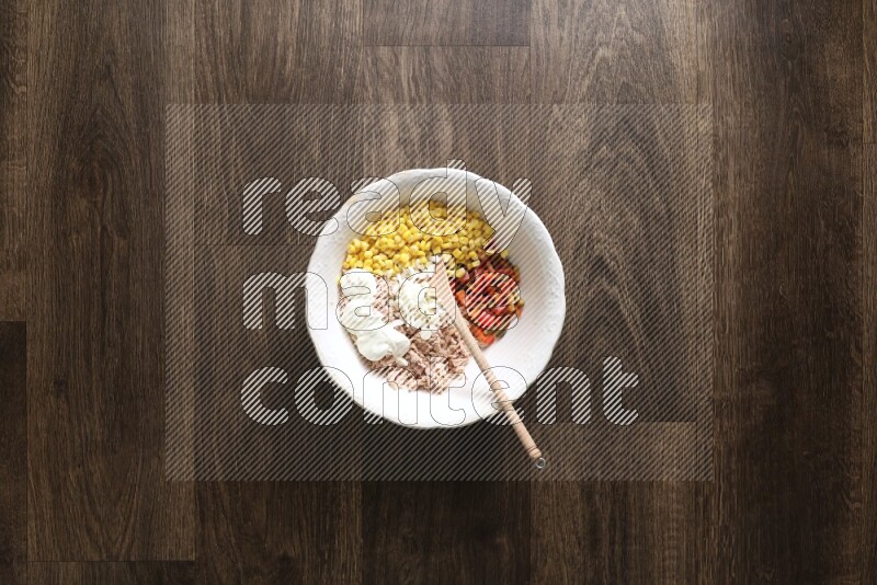 A white bowl full of tuna, colored bell pepper, sour cream, corn, parsley, black pepper powder and sauce, with wooden spoon on wooden background