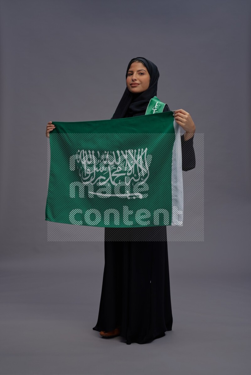 Saudi woman wearing Abaya standing holding Saudi flag on gray background