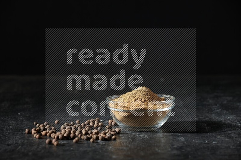 A glass bowl full of allspice powder and whole balls spreaded on a black flooring