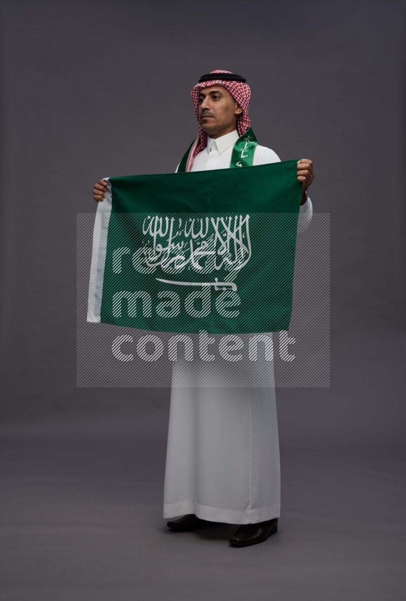 Saudi man wearing thob and shomag standing holding Saudi flag on gray background