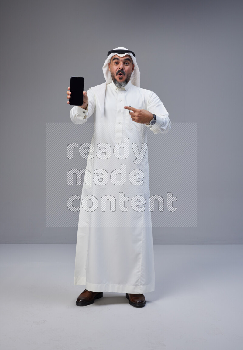 Saudi man Wearing Thob and white Shomag standing showing phone to camera on Gray background