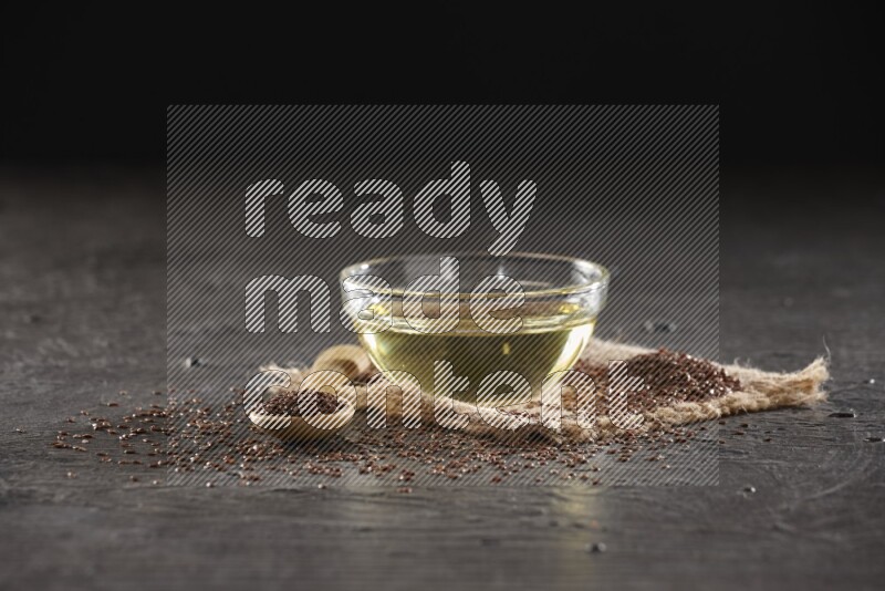 وعاء زجاجي ممتلئ بزيت بذر الكتان مع ملعقة خشبية ممتلئة بحبوب بذر الكتان مع بعض الحبوب المتناثرة علي قطعة من القماش علي خلفية سوداء