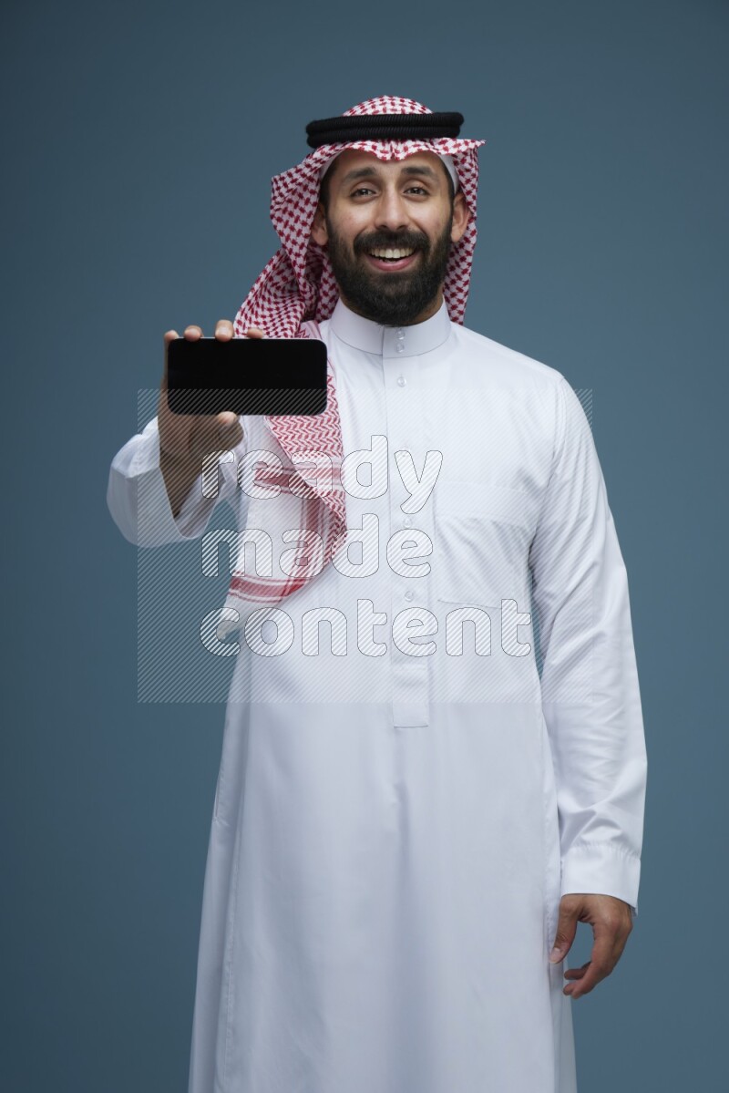 A man showing his phone Screen in a blue background wearing Saudi Thob with Shomag