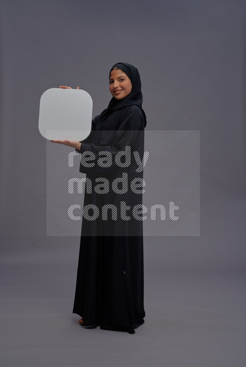 Saudi woman wearing Abaya standing holding social media sign on gray background