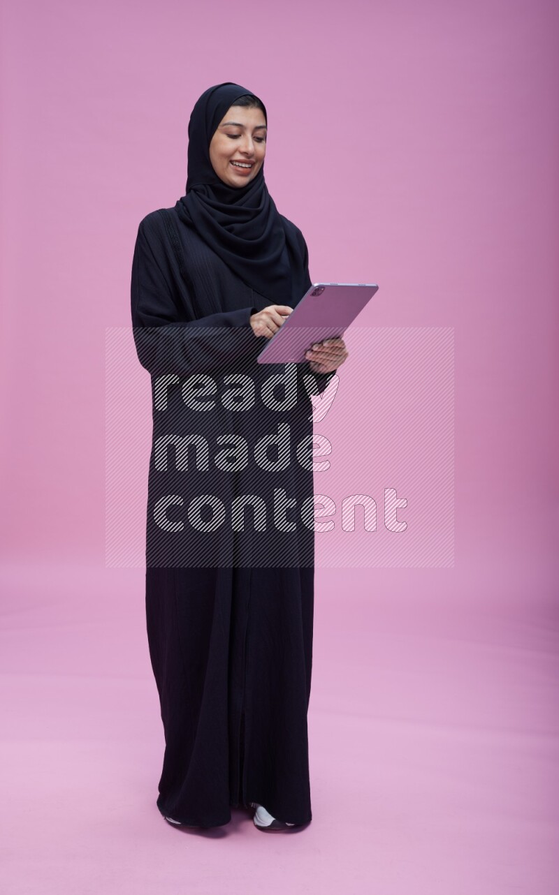 Saudi woman wearing Abaya standing working on tablet on pink background