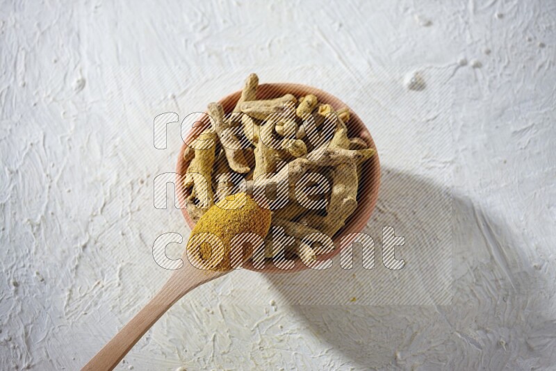 A wooden spoon full of turmeric powder above a wooden bowl full of dried turmeric whole fingers on a textured white flooring