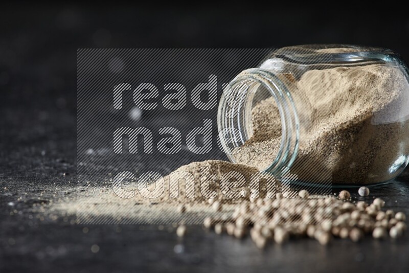 وعاء زجاجي مقلوب ممتلئ ببودرة الفلفل الأبيض مع بودرة وحبوب فلفل متناثرة على أرضية سوداء