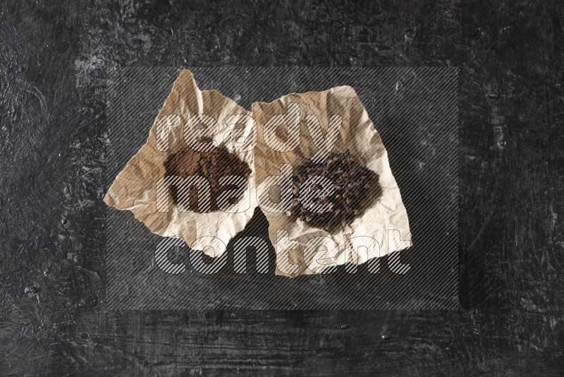 Cloves powder and cloves in 2 crumpled pieces of paper on a textured black flooring