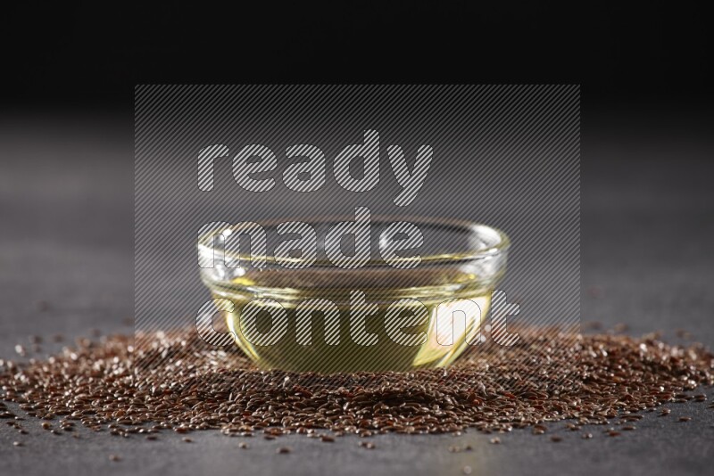 A glass bowl full of flaxseeds oil surrounded by the seeds on a black flooring
