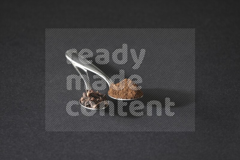 2 metal spoons, one full of cloves grains and the other full of cloves powder on a black flooring