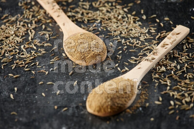 2 wooden spoons full of cumin powder with spreaded seeds on textured black flooring