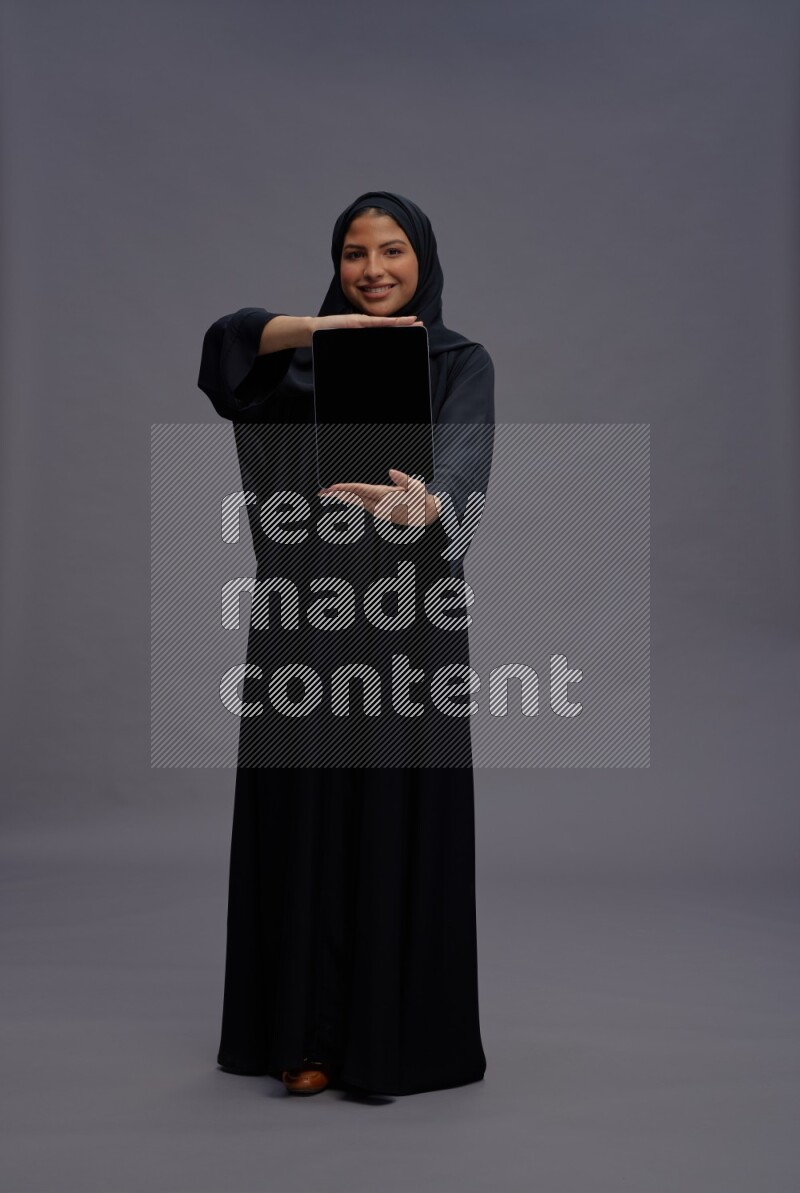 Saudi woman wearing Abaya standing showing tablet to camera on gray background