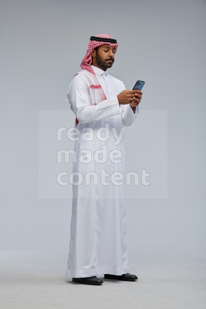 Saudi man Wearing Thob and shomag standing texting on phone on Gray background