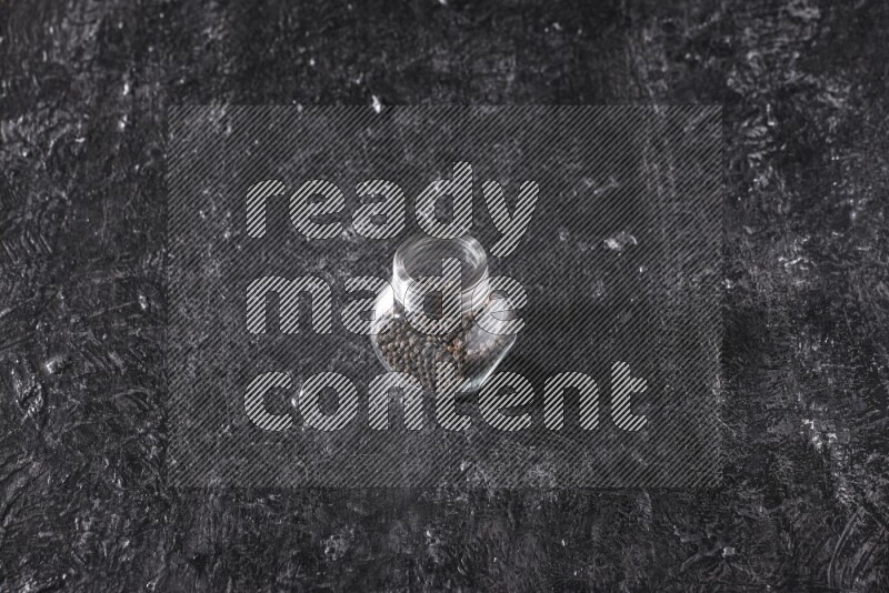 A glass spice jar full of black pepper beads on textured black flooring