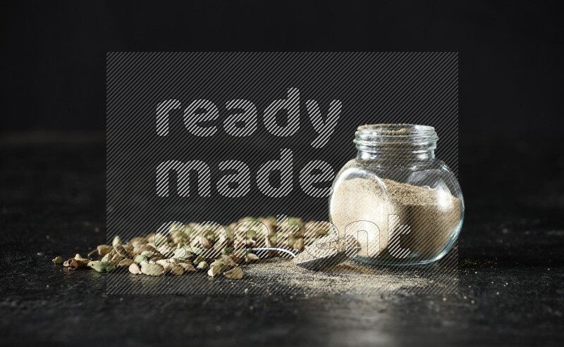 A glass spice jar and a metal spoon full of cardamom powder and cardamom seeds spreaded on textured black flooring