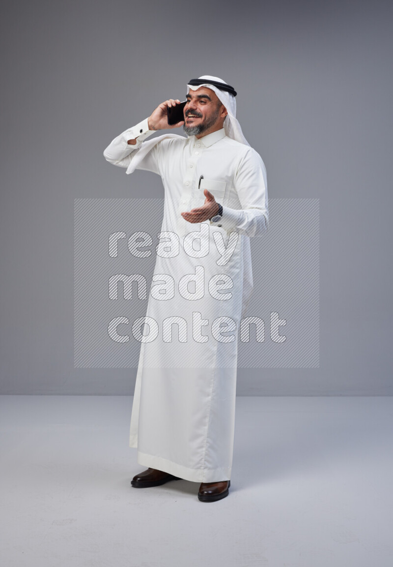 Saudi man Wearing Thob and white Shomag standing talking on phone on Gray background