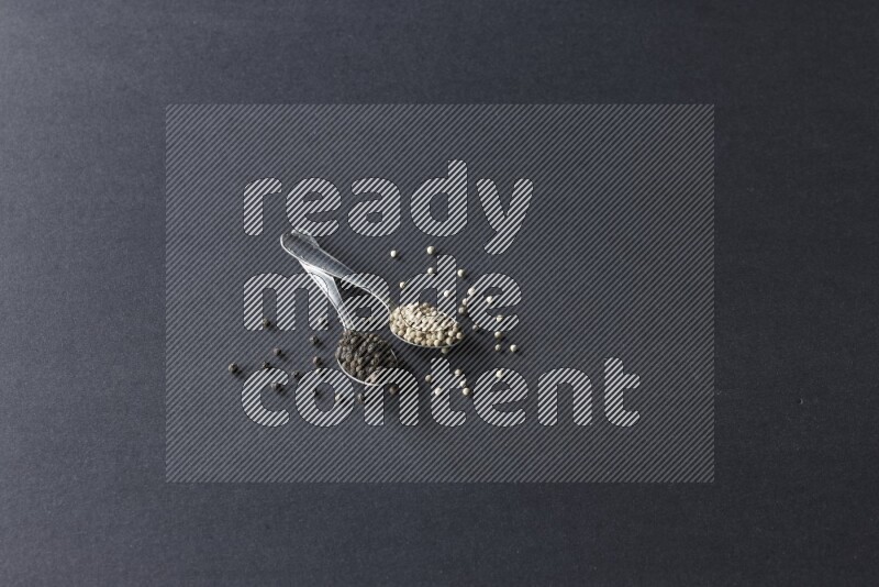 2 metal spoons full of black and white pepper beads with spreaded beads on black flooring