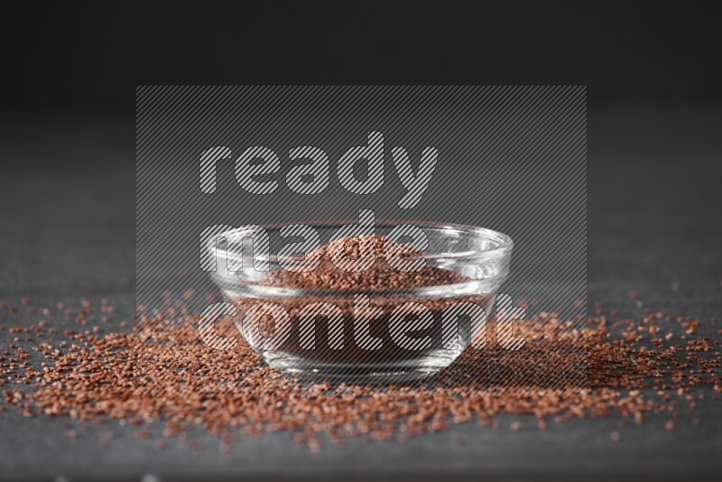 A glass bowl full of garden cress seeds surrounded by seeds on a black flooring