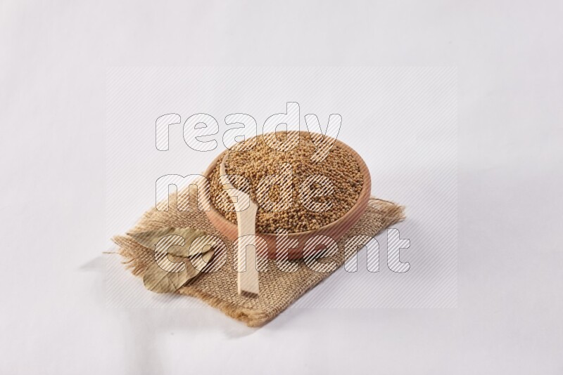 A wooden bowl full of mustard seeds with a wooden spoon on the seed set on burlap fabric on a white flooring